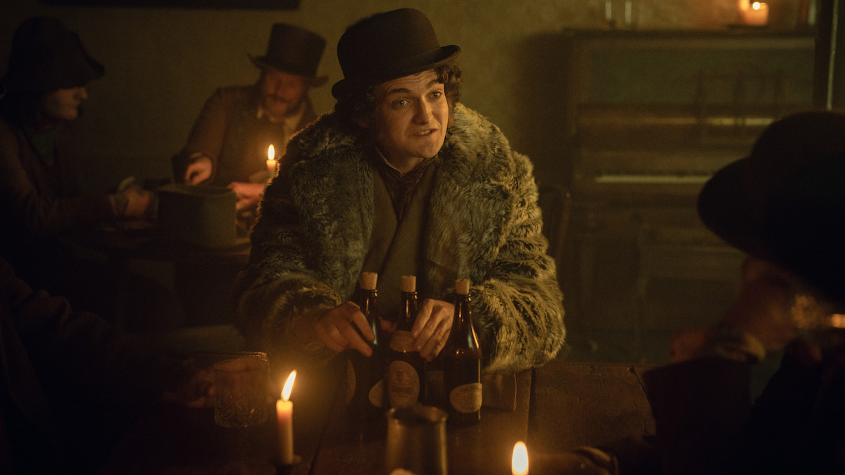 Jack Gleeson as Byron Hedges wears a fur coat and holds bottles of beer in ‘House of Guinness’ Season 1.