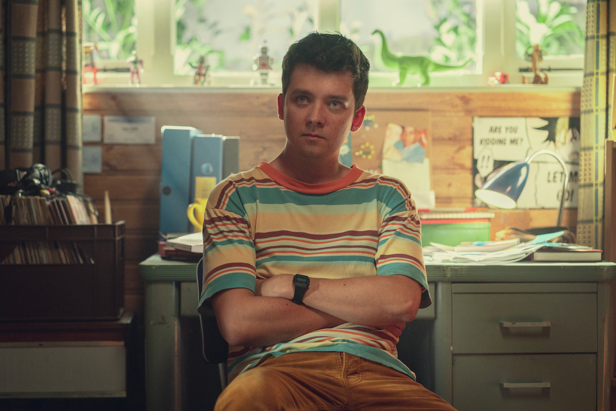 Asa Butterfield as Otis Milburn sits at a desk with his arms folded in Season 4 of ‘Sex Education’