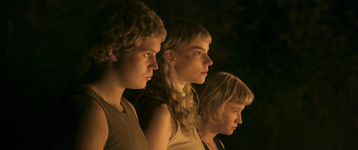 Three people with short blonde hair stand side by side in dim, warm light, looking intently ahead; they appear to be outdoors at night, illuminated by a soft, glowing source.