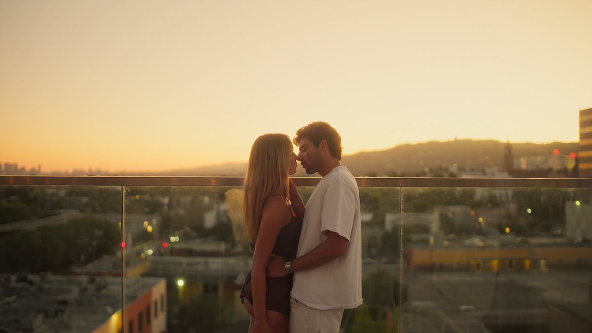 A couple stands close together on a rooftop at sunset, with city buildings and hills in the background, sharing an intimate moment.