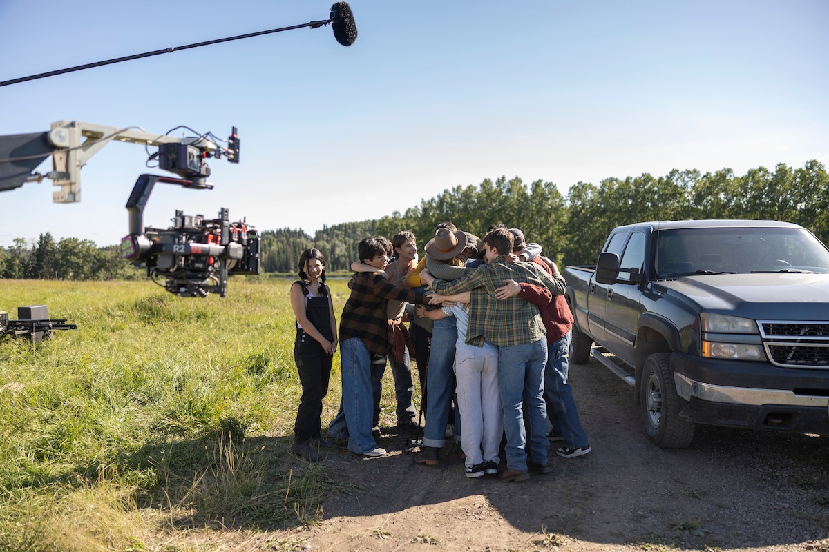A group of people stands in a circle hugging in a grassy field beside a pickup truck, with a film crew and camera crane visible, suggesting a movie or TV shoot outdoors on a sunny day.
