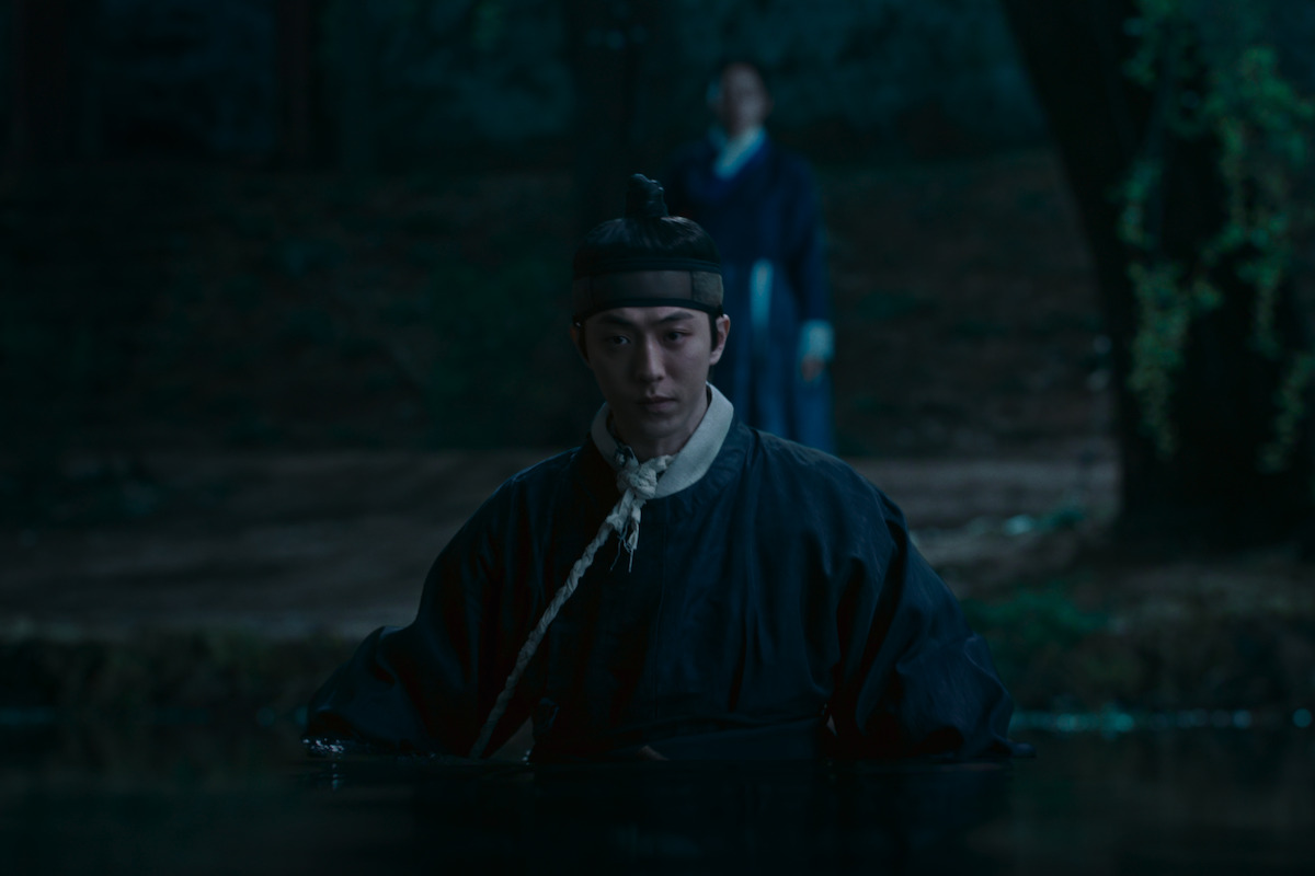 A man in traditional Korean clothing stands in water at night, with another person blurred in the background in a forested environment. The setting is dark and atmospheric, suggesting a dramatic or tense moment.