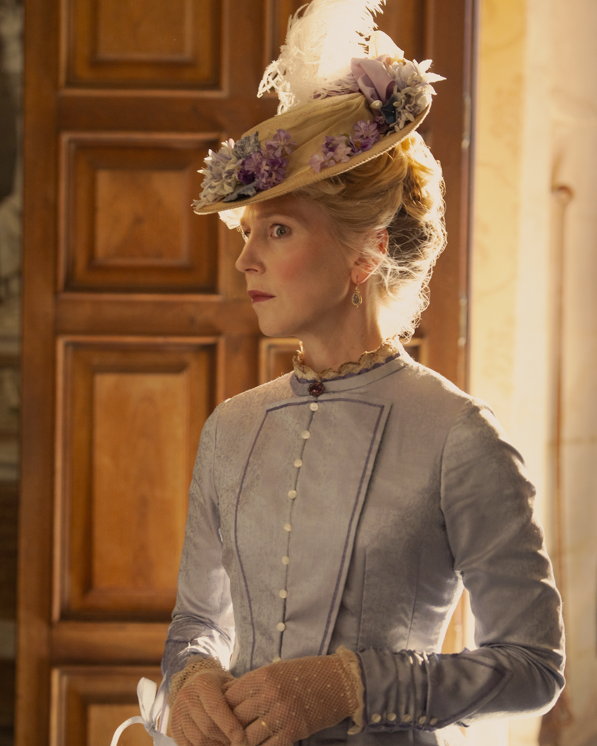 A woman in Victorian-era attire stands indoors, wearing a light blue dress with pearl buttons and a decorative hat adorned with flowers, in front of a wooden door and sunlit wallpapered wall.