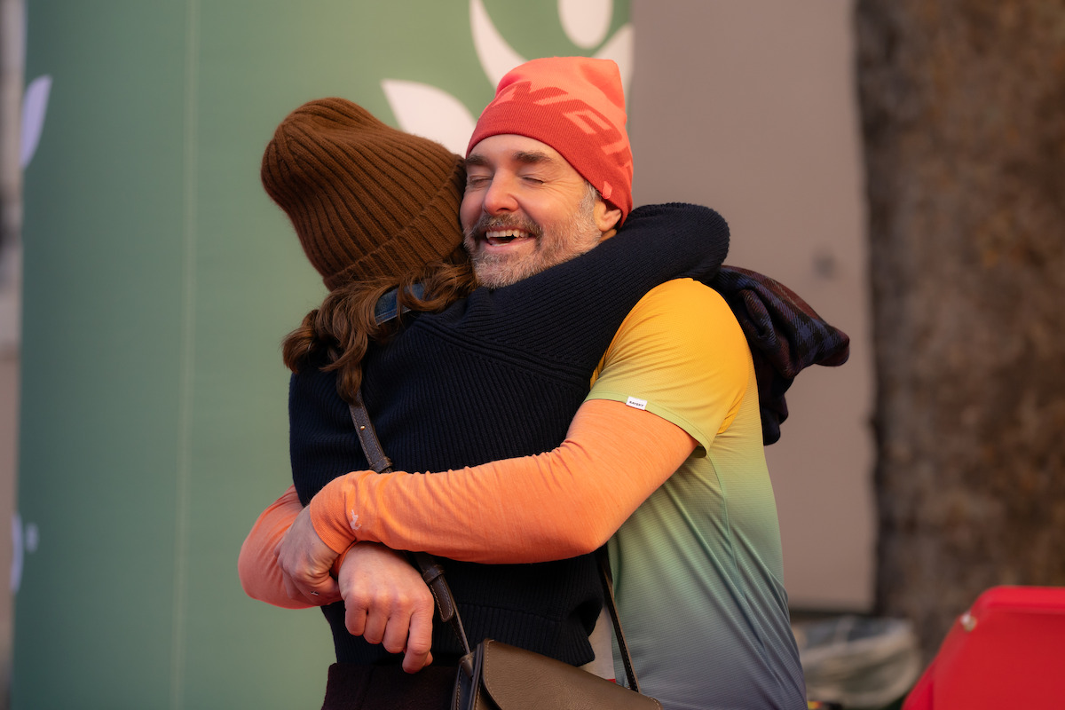 Two people dressed in winter clothing hugging outdoors, with one person smiling warmly. The background features a green wall with a logo and a tree trunk, suggesting a friendly or festive public setting.