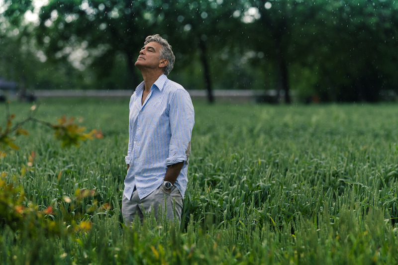 Jay Kelly (George Clooney) stands in a grassy field in the countryside of Italy wearing a button up shirt with head titled to the sky, eyes closed, as rain falls from the sky.