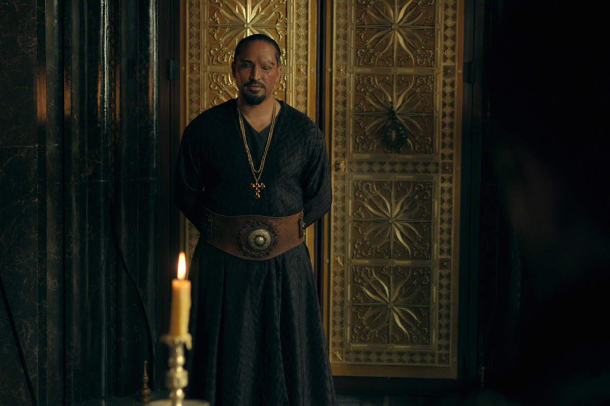 A man in dark medieval-style clothing stands in a dimly lit, ornate room with golden doors behind him, exuding a solemn and serious mood. A lit candle is visible in the foreground, adding to the atmosphere.