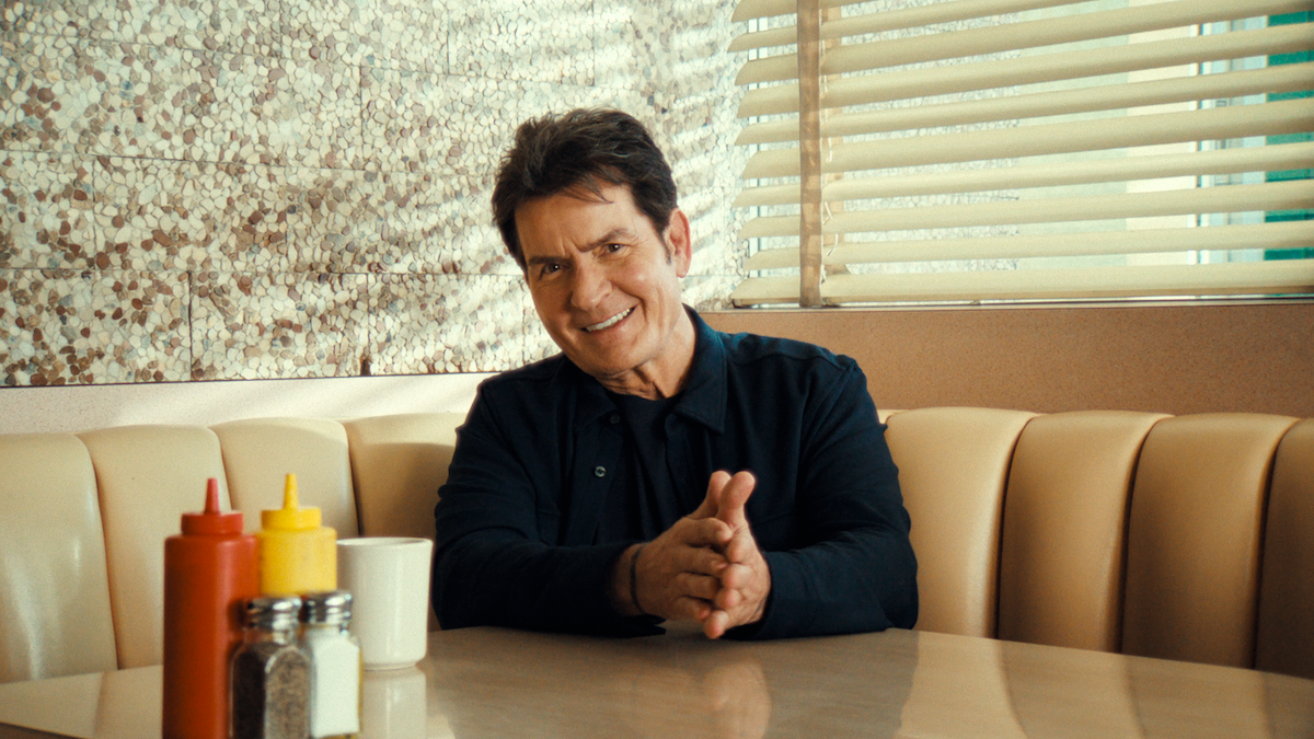 Man smiling while sitting in a retro diner booth, with sunlight streaming through venetian blinds, condiments, and a coffee mug on the table.