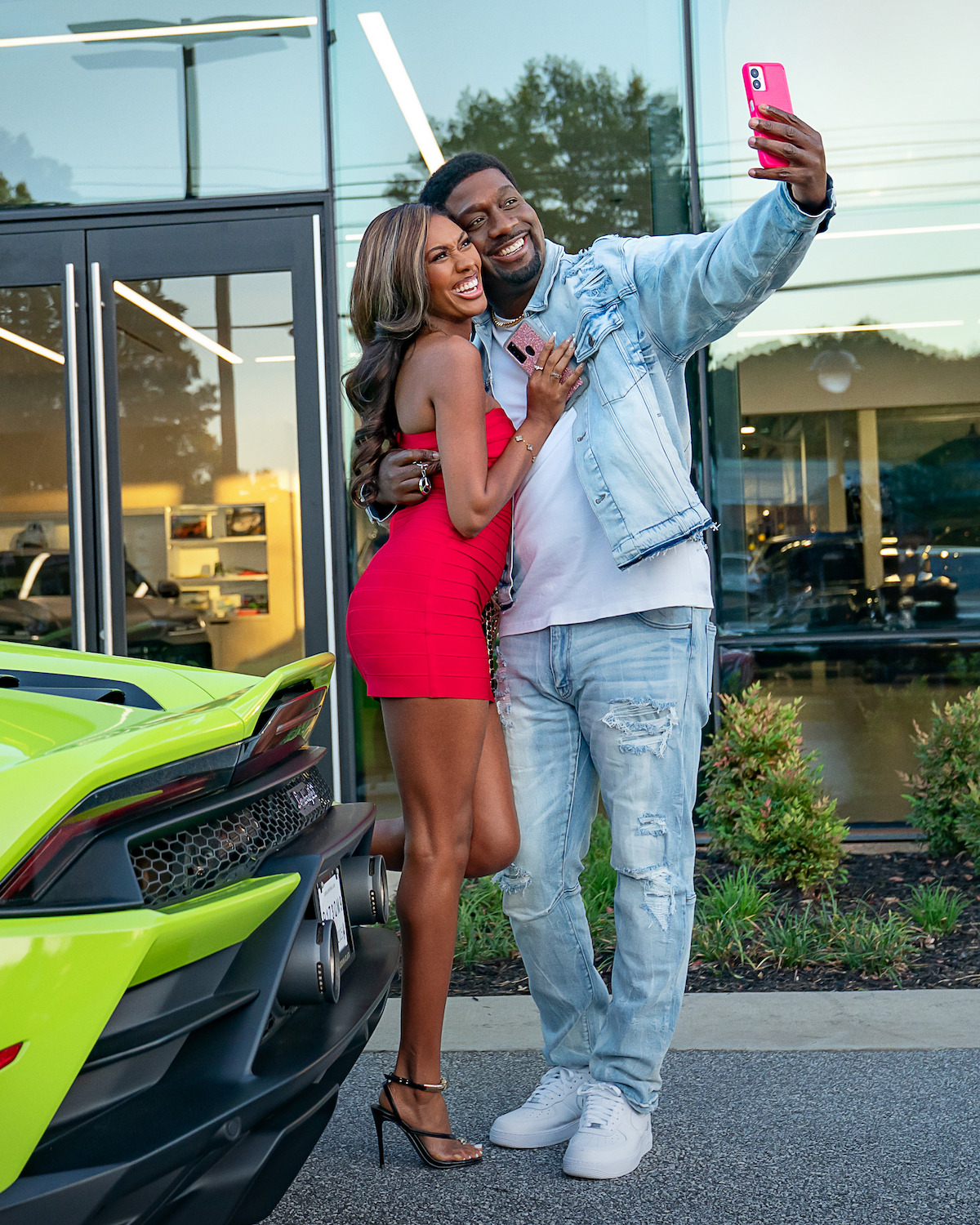 A smiling couple poses for a selfie beside a bright green sports car outside a modern building with glass windows, surrounded by greenery and reflections of trees.