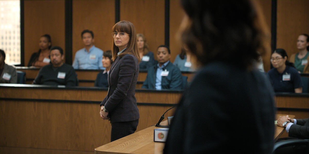 A woman in professional attire stands in a courtroom before a jury, facing another person in the foreground, with several jurors seated in the background observing the proceedings.