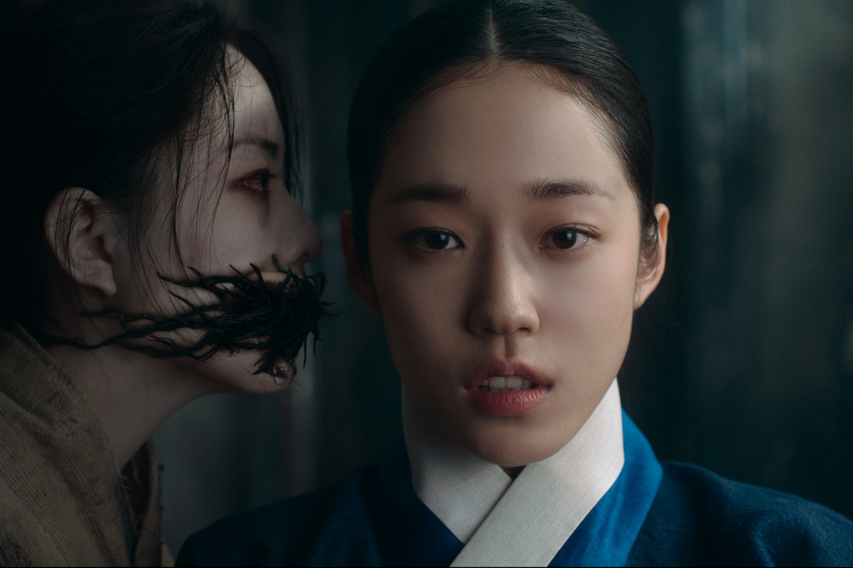 Two women in traditional Korean attire, one with a fearful expression in the foreground and another with a disfigured, dark, vine-like mouth whispering behind her in a dimly lit, tense setting.