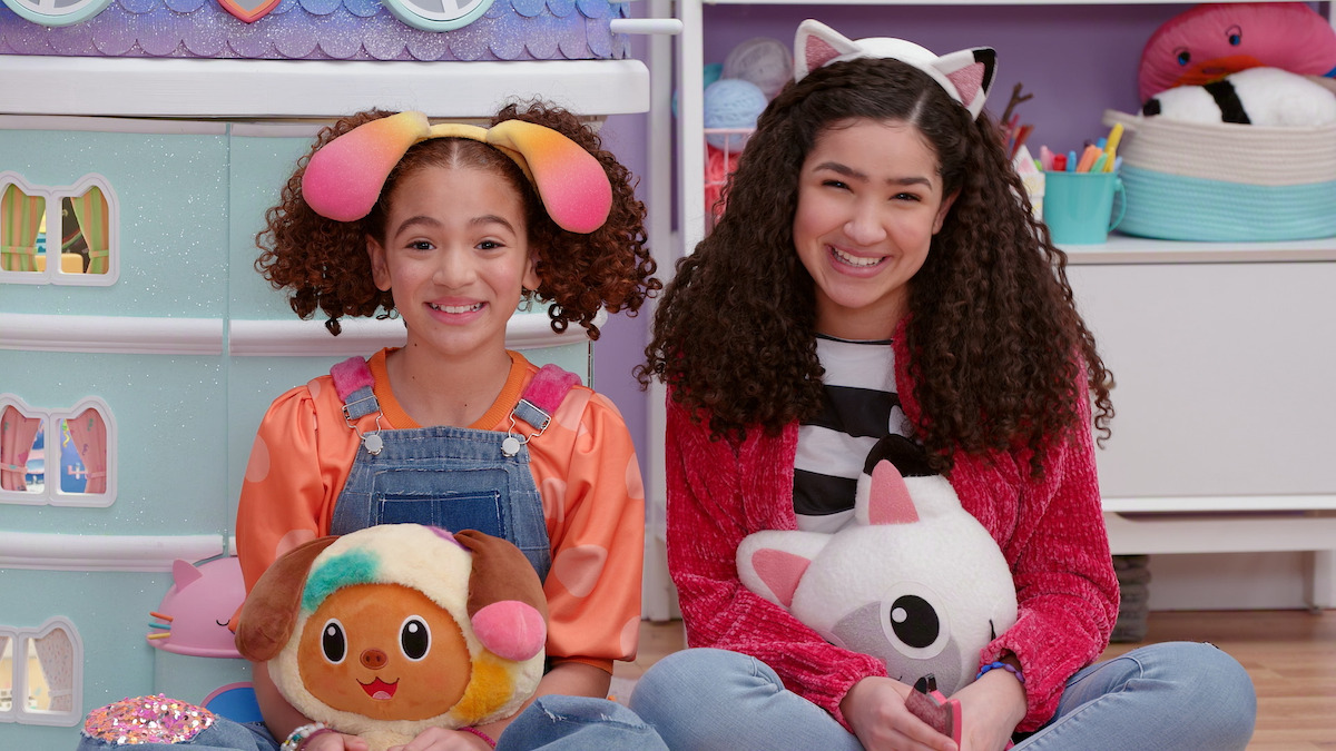 Two smiling girls sit on the floor in a colorful playroom, holding plush toys. One wears dog ear headband and overalls, the other has a cat ear headband and red jacket. Shelves and art supplies are visible in the background.