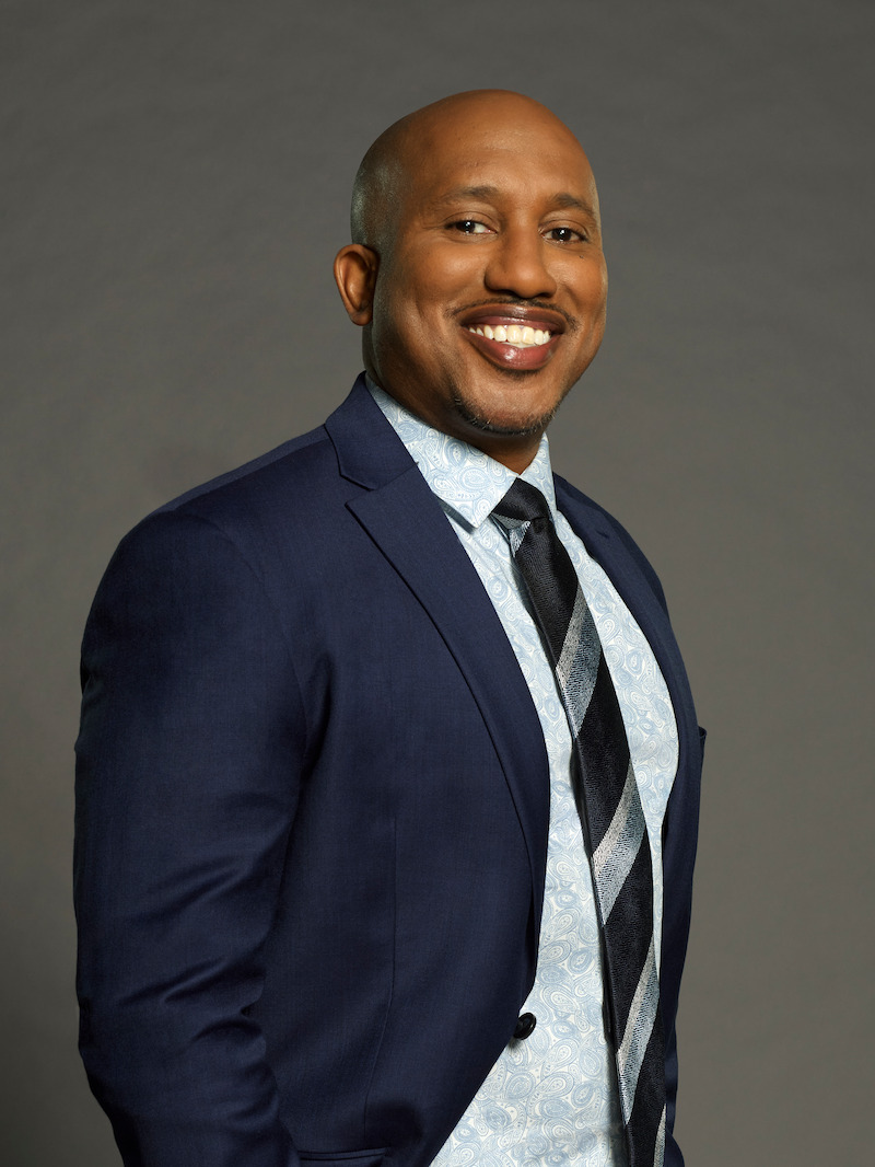Smiling man in a blue suit, patterned shirt, and striped tie poses against a plain grey studio background, exuding professionalism and confidence.