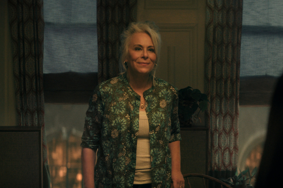 Jane Kaczmarek as Lilly Cooper with light hair smiling, standing in a warmly lit room with patterned curtains, two windows, and plants in the background, wearing a green floral shirt over a light top.