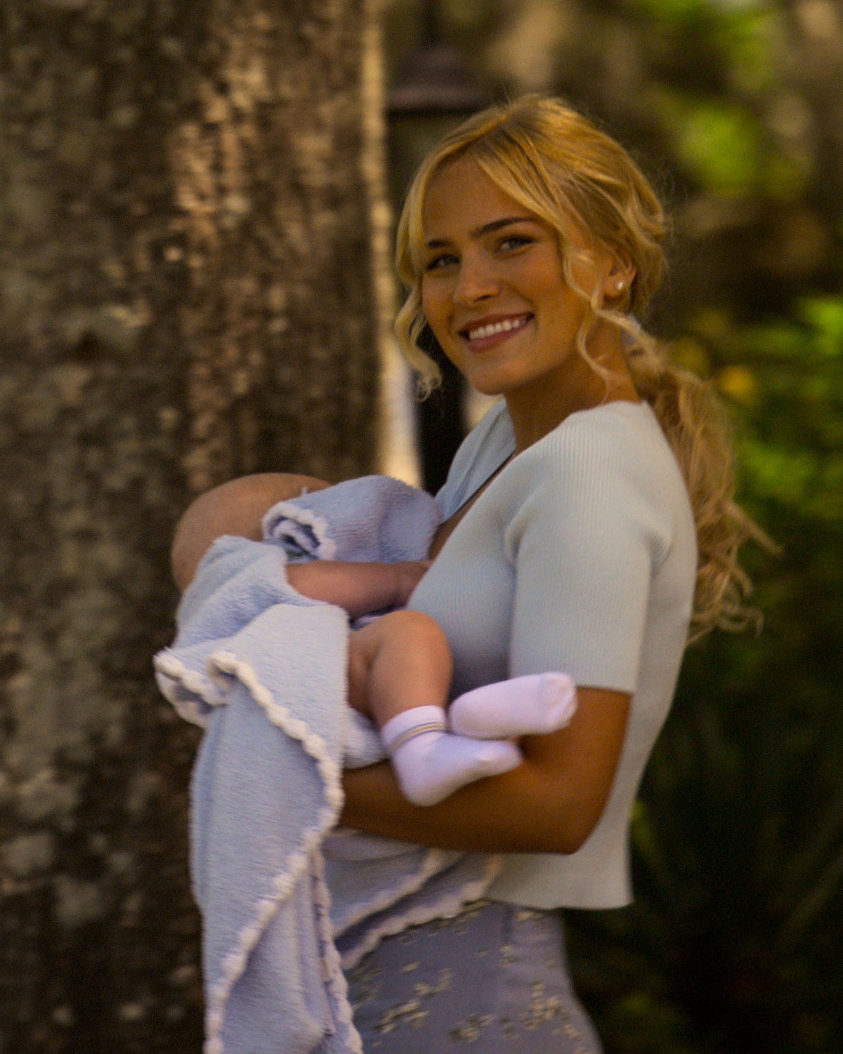 Tommi Rose as Larissa Genrette, holding a baby wrapped in a light blue blanket outdoors near a tree, with greenery and soft sunlight in the background.