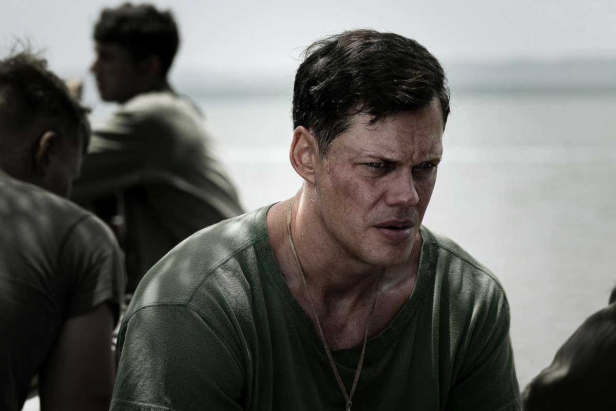 Bill Skarsgard as Bob Baumann in a military-style green shirt sits outdoors near water, looking serious and thoughtful, with other men in similar attire blurred in the background suggesting a somber, reflective scene.