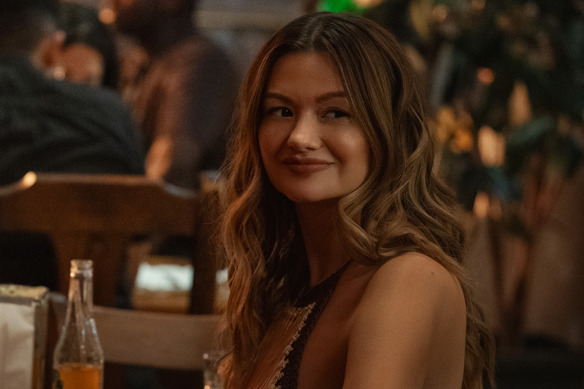 A young woman with wavy hair sits at a wooden table in a warmly lit restaurant, smiling slightly, with a bottle of soda on the table and other diners blurred in the background.