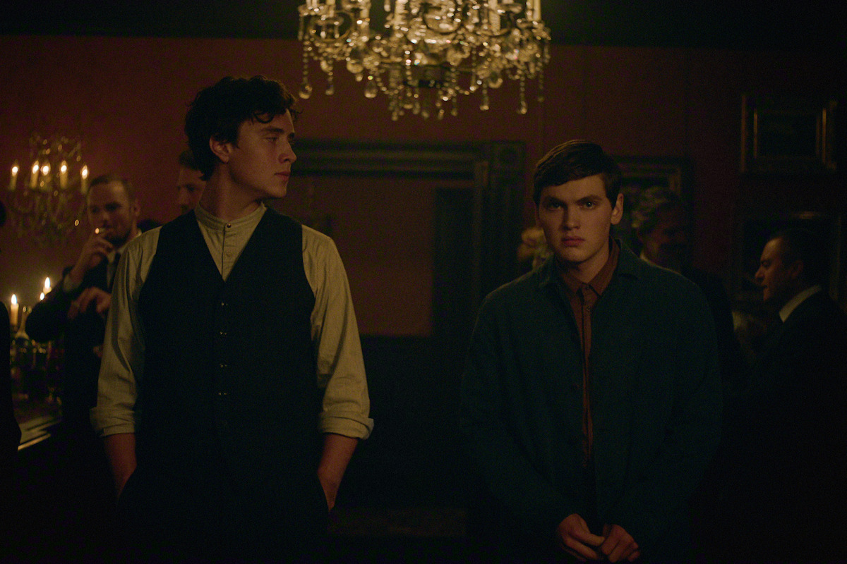 Two young men stand in a dimly lit, elegant room with a chandelier and candelabras, dressed in vintage-style clothing, surrounded by other people engaged in conversation.