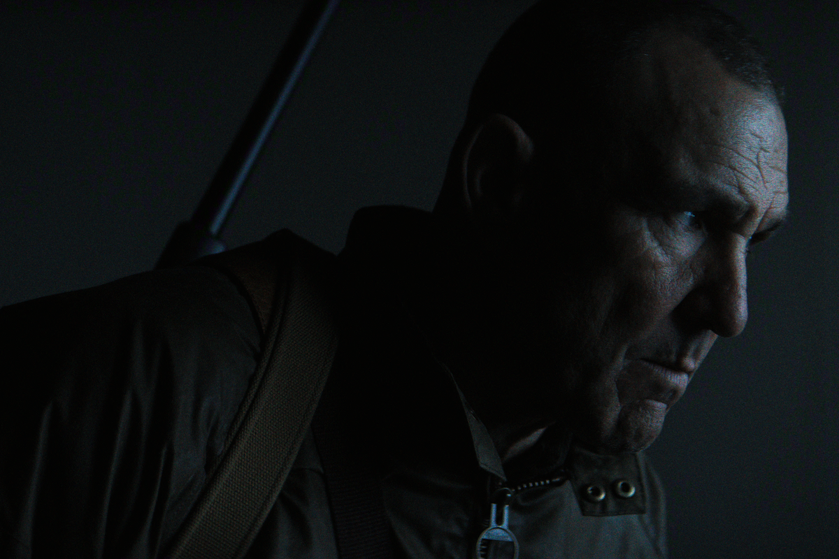 A man with a serious expression is in a dimly lit setting, wearing a brown jacket and carrying a strap over his shoulder, suggesting a tense or dramatic atmosphere.