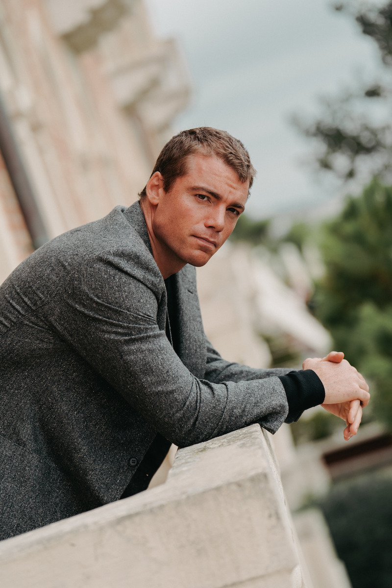 Gabriel Basso in a gray suit jacket stands on a balcony in Venice.