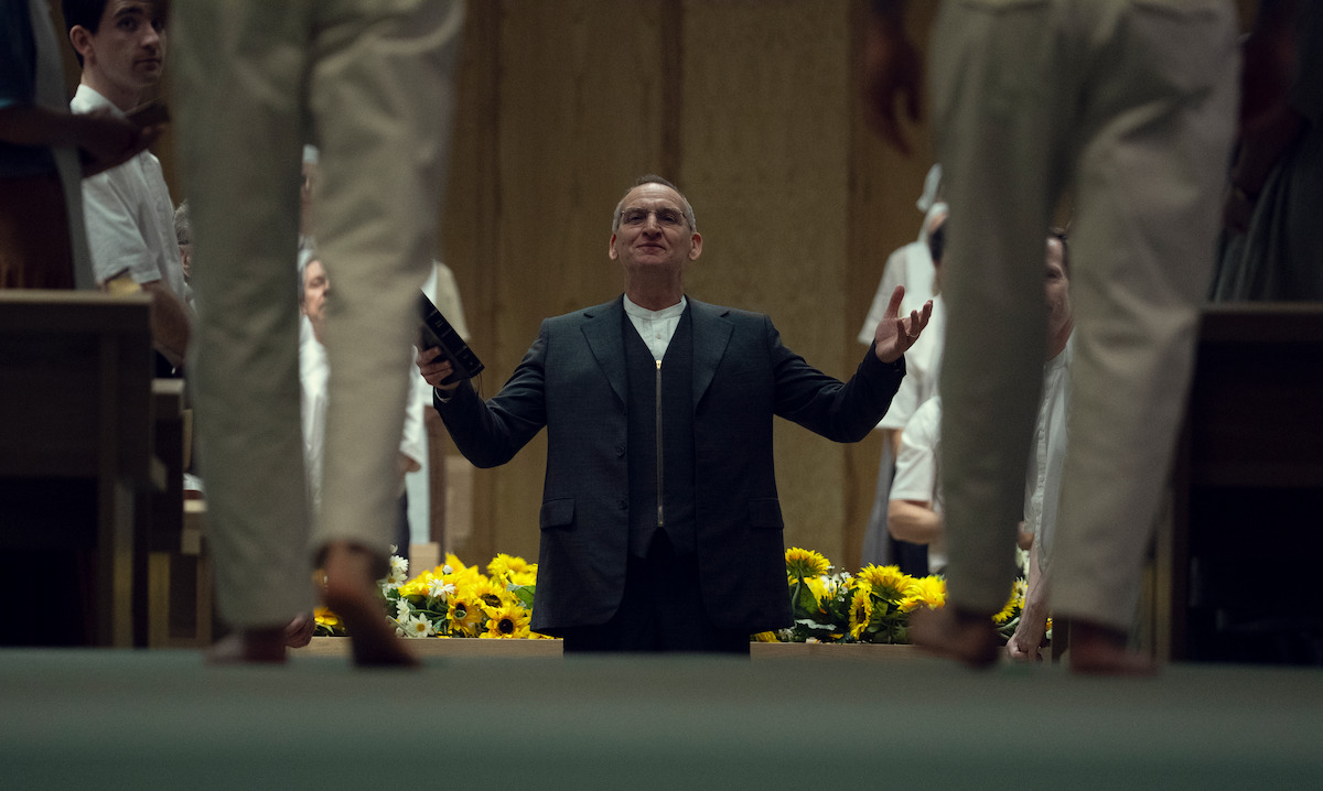 A man in a suit stands confidently with arms outstretched, holding a book, in front of people dressed in white in an indoor setting with yellow flowers and wooden panels, creating a solemn or ceremonial atmosphere.