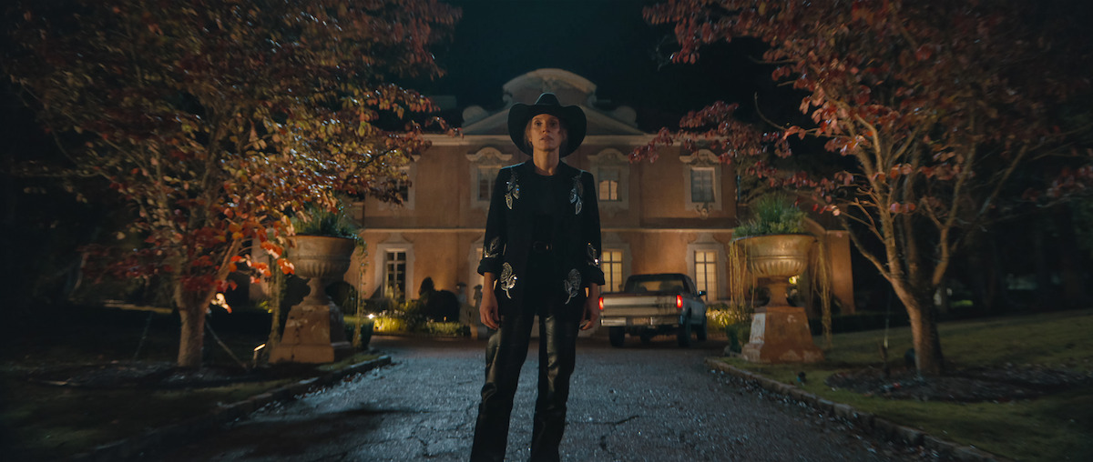 Person in a black cowboy outfit stands on a driveway at night, with autumn trees on both sides and a large, well-lit house and parked cars in the background.