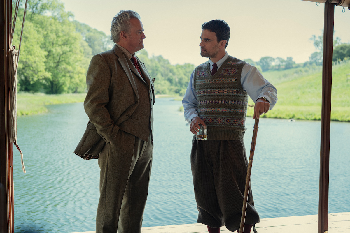 Two men in aristocratic clothing talk by a riverbank; one holds a drink, the other a walking stick. They stand under a shaded structure with green trees and calm water visible in the background on a sunny day.