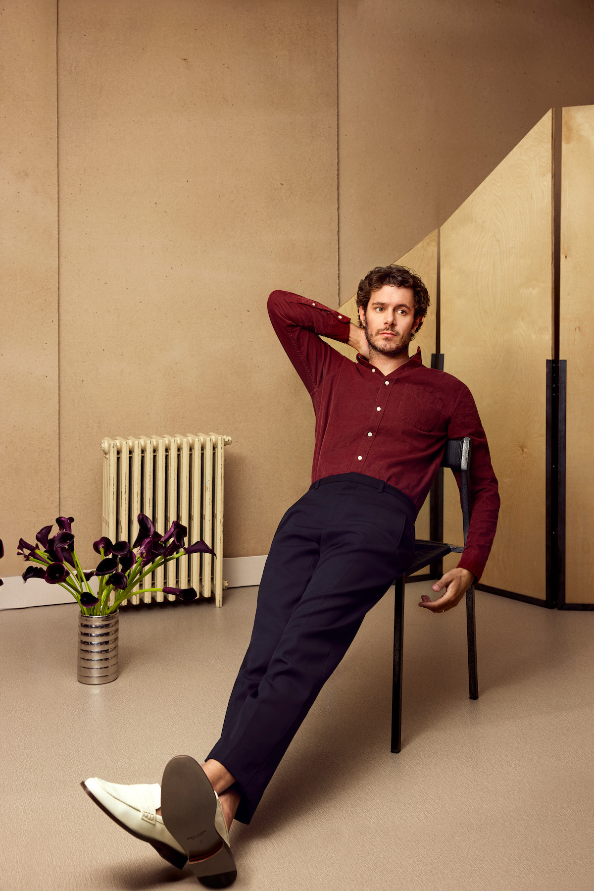 Adam Brody lounges on a chair wearing a maroon button up shirt and black pants