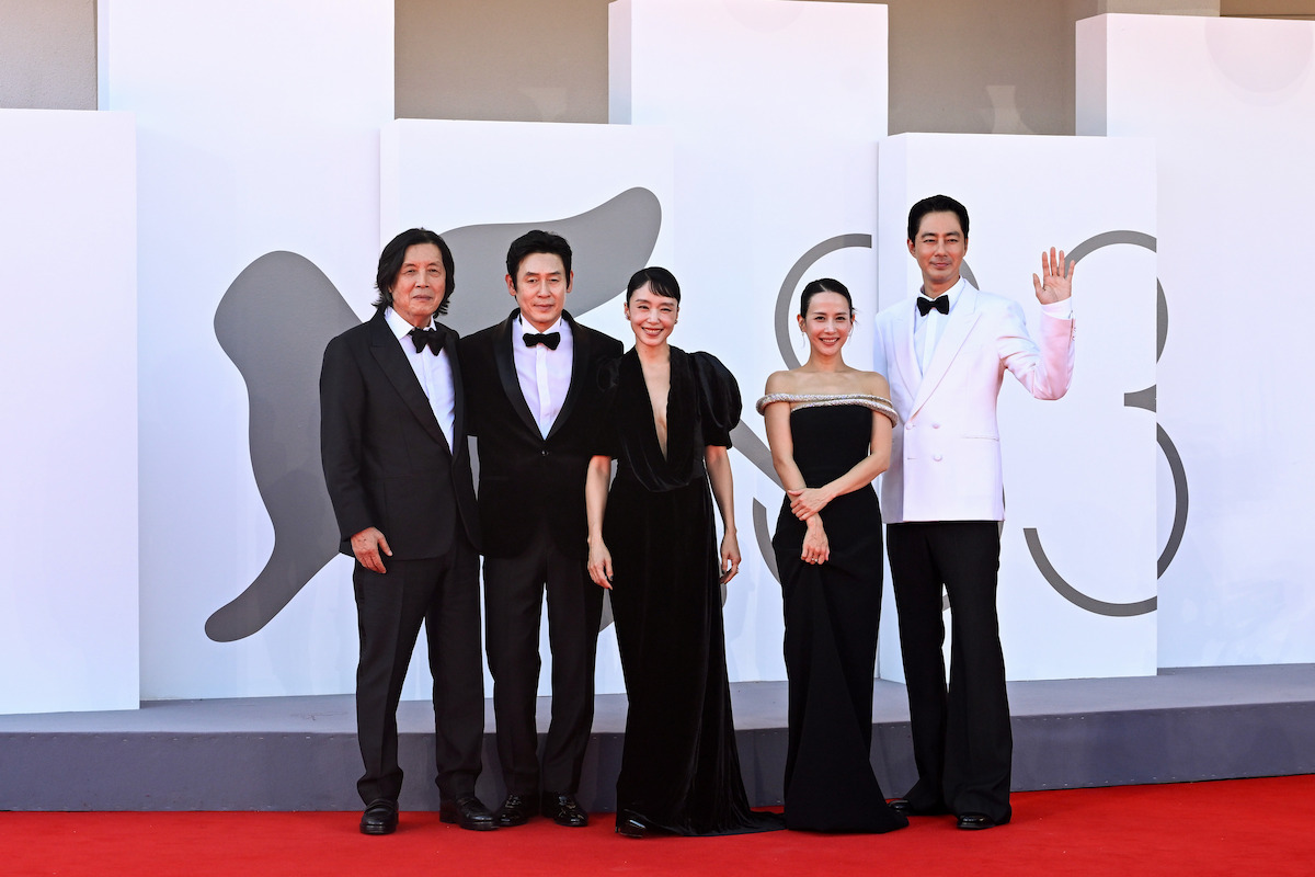 Five people dressed formally pose together on a red carpet at an event with a white backdrop and abstract designs, suggesting a film festival or awards ceremony setting. One person waves while others smile at the camera.