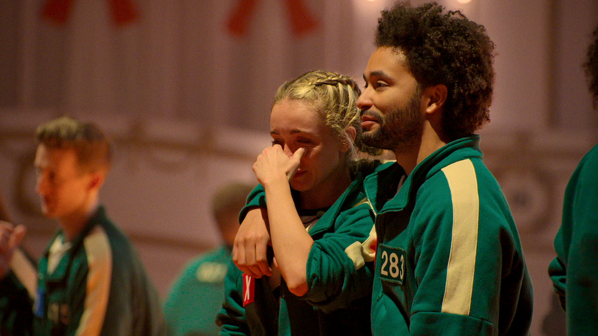 Two people in matching green tracksuits stand close together indoors, one comforting the other who appears emotional, with blurred people in the background. The mood is tense and supportive.