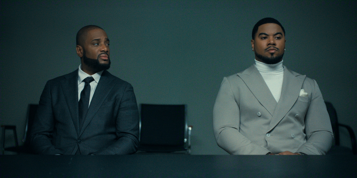 Two well-dressed men sit at a table in a dimly lit, modern room with empty black chairs behind them, suggesting a formal or professional setting, possibly an interview or meeting.