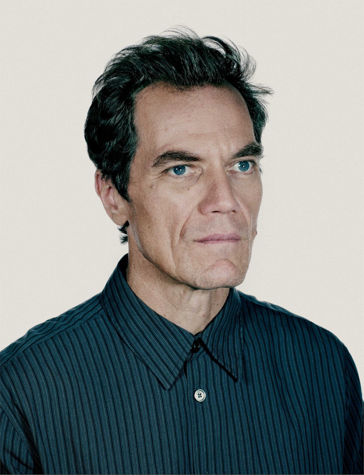 Middle-aged man in a striped button-up shirt looking slightly upward with a serious expression, isolated against a plain light background, evoking a neutral or contemplative mood.