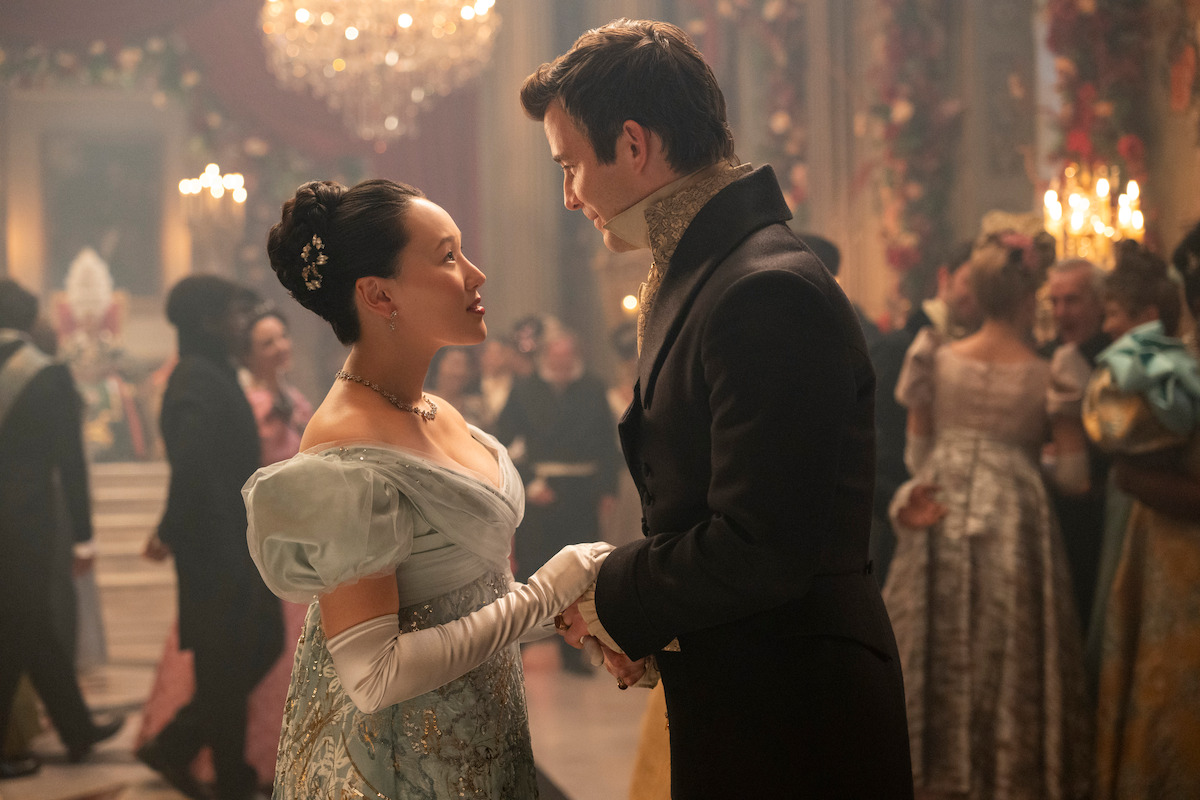 A woman and man in elegant Regency-era attire share a dance and gaze at each other in a lavish ballroom filled with other guests, chandeliers, and ornate decorations, evoking a formal historic setting.