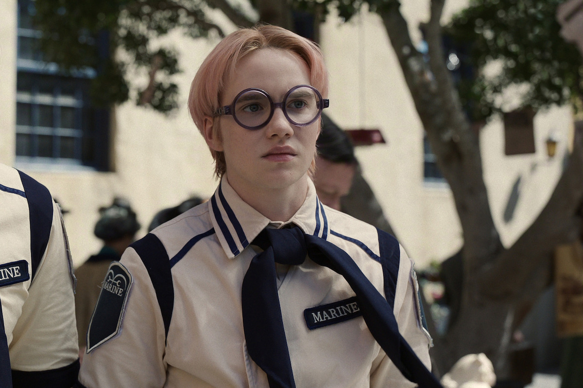 Person with short pink hair and round glasses wearing a cream and navy uniform with "Marine" patch, standing outdoors in a shaded area with trees and a building in the background.