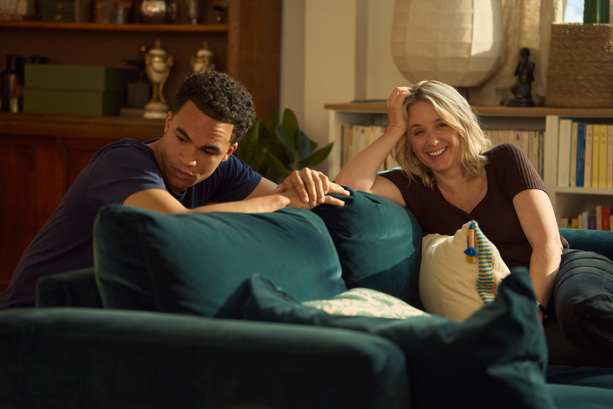 Two people sitting on a teal sofa in a cozy living room. The man looks thoughtful while the woman smiles warmly. Shelves with books, plants, and decor are in the background, creating a homey atmosphere with soft natural light.