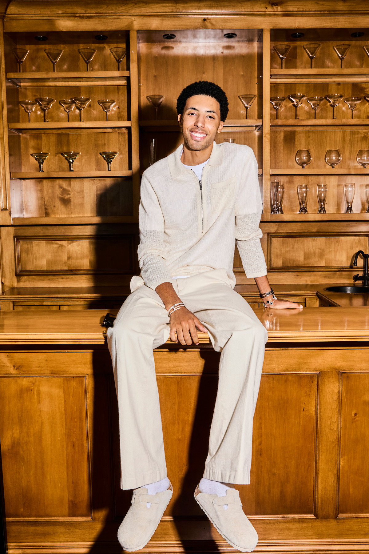 Smiling man in light-colored clothing sitting on a polished wooden bar in a room with shelves of various glasses behind him, creating a relaxed and elegant atmosphere.