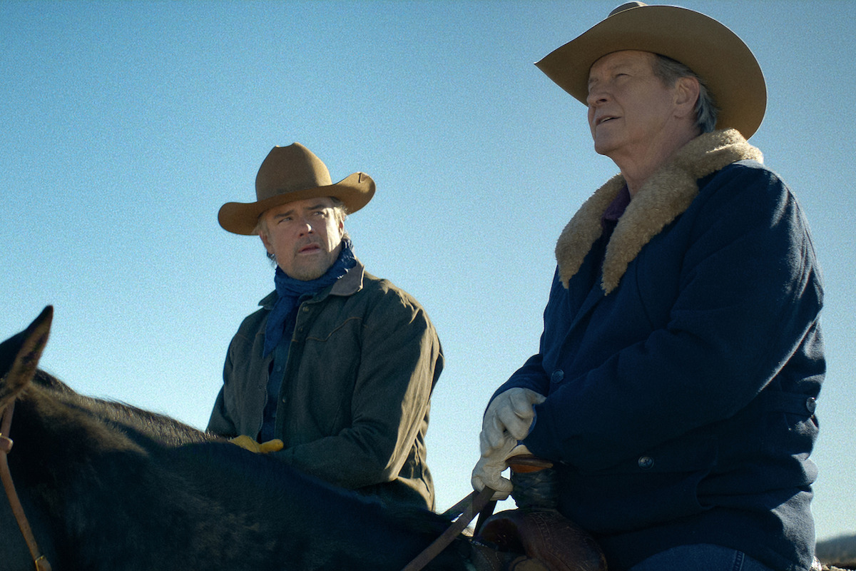 Two men wearing cowboy hats and western jackets sit on horseback outdoors under a clear blue sky, appearing to converse in a rural or ranch setting.