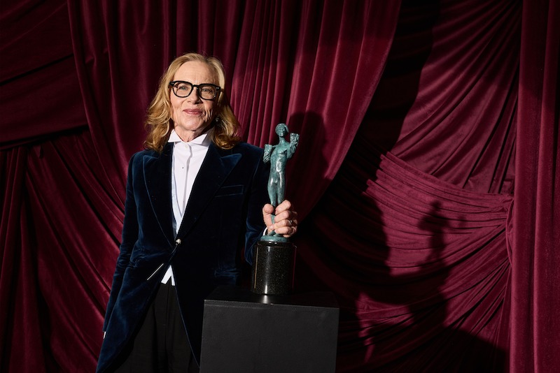 Amy Madigan in formal attire holding a statue award, standing in front of rich red velvet draped curtains, under stage lighting.