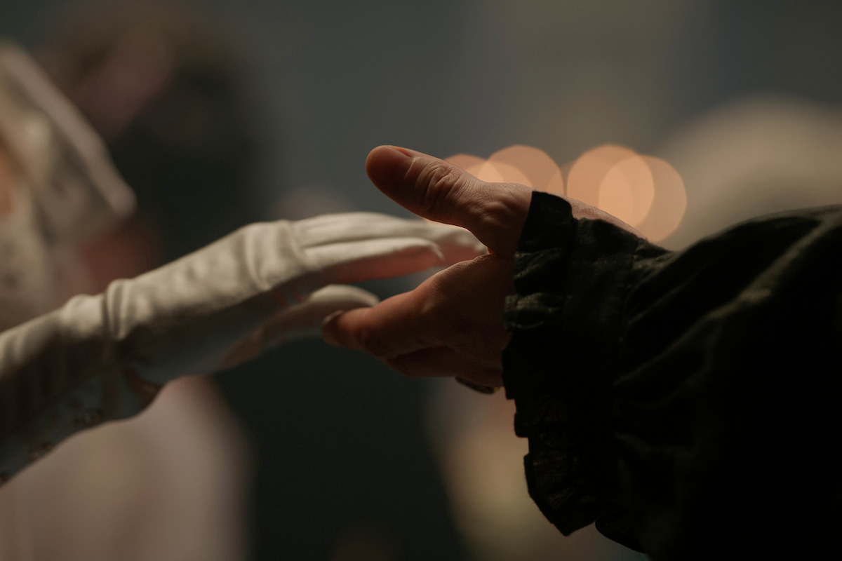 A gloved hand reaches for an ungloved hand dressed in formal dark attire, in a dimly lit, elegant setting, suggesting a historical or romantic event.