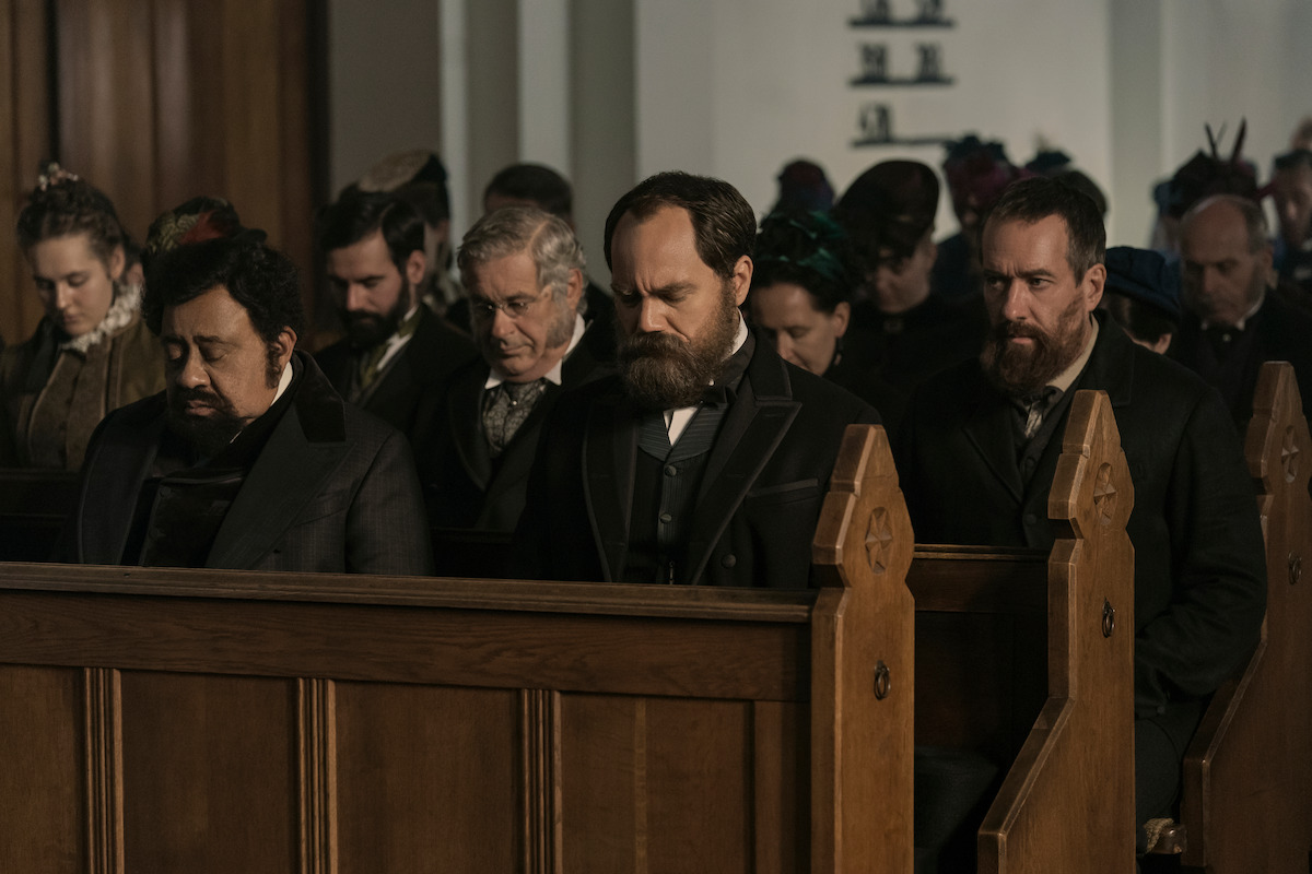 Men and women in period clothing sit in wooden pews inside a church, heads bowed in solemn prayer. The lighting is subdued, evoking a somber and reflective mood. The setting is historical and reverent.