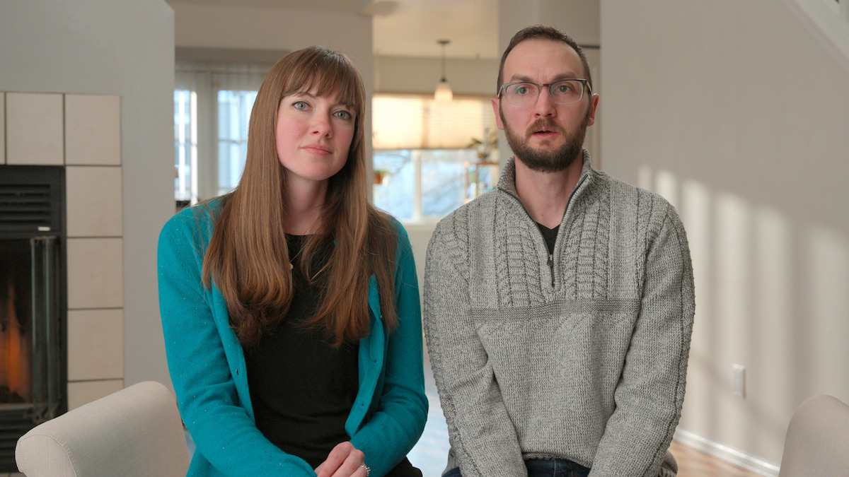 Two adults sitting indoors on chairs in a bright living room, with a fireplace on the left and natural light from large windows in the background.