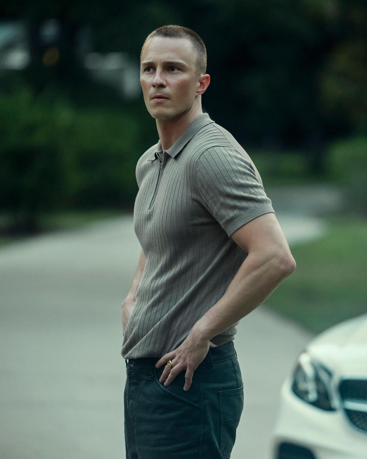 Drew Starkey as Rafe Cameron, standing outdoors on a street near a white car, wearing a light brown short sleeve polo shirt and dark pants, with hands in pockets and a serious expression, greenery and trees blurred in the background.