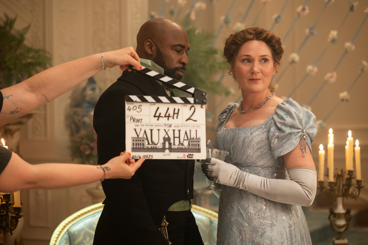 Actors in period costumes on a film set, one woman in a blue gown and gloves, one man in dark attire, a clapboard is being held in front of them, ornate room with candles and floral decorations in the background.