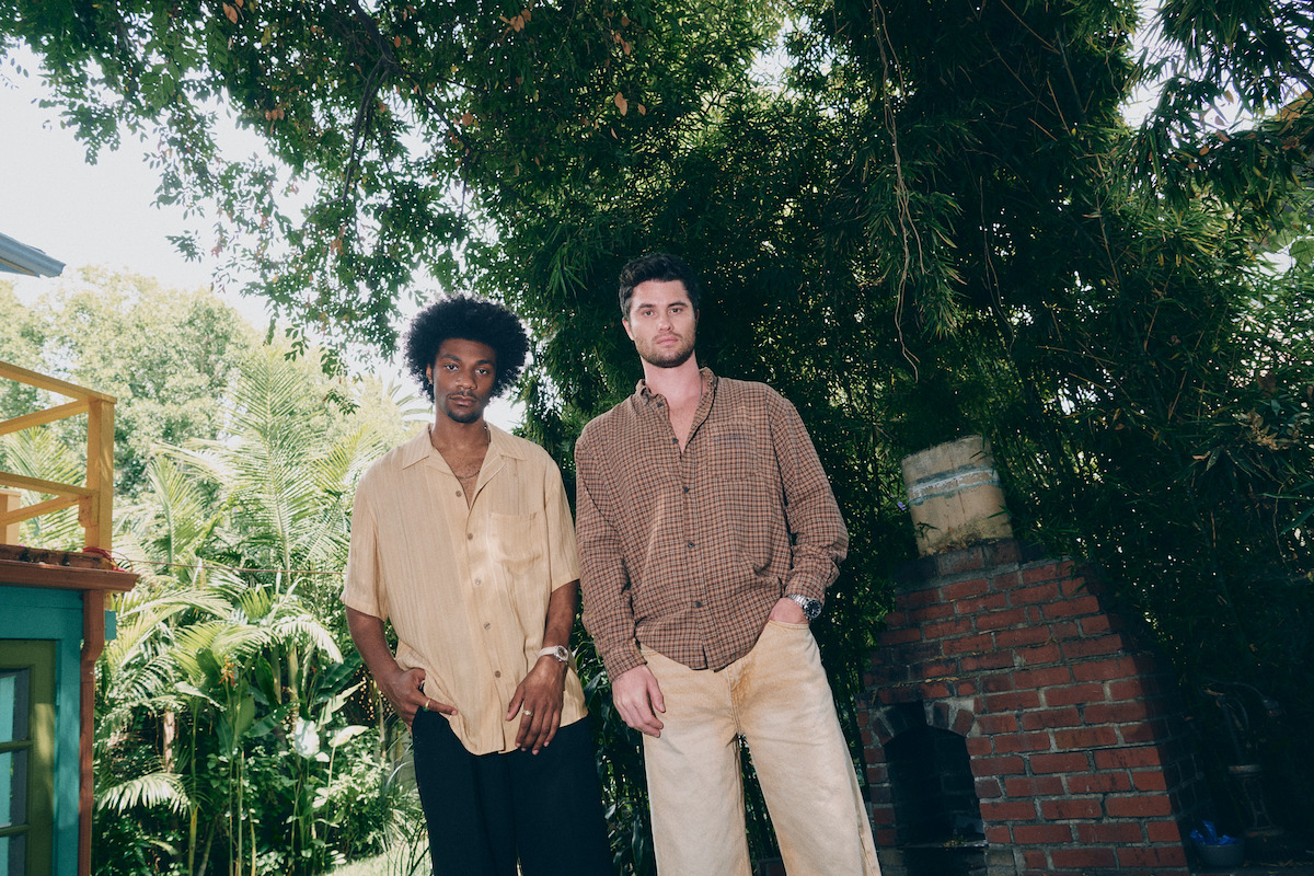 Two men stand outdoors under a large tree, next to a brick structure, with lush greenery and sunlight filtering through the leaves in a relaxed, casual setting.