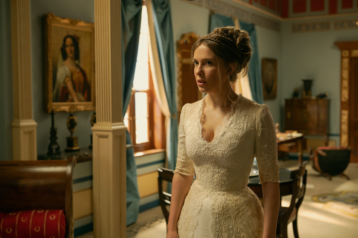Woman in an elaborate white gown and large feathered headdress stands in an ornate, gold-trimmed room, surrounded by three women dressed in dark period clothing, as they adjust her outfit and admire her.