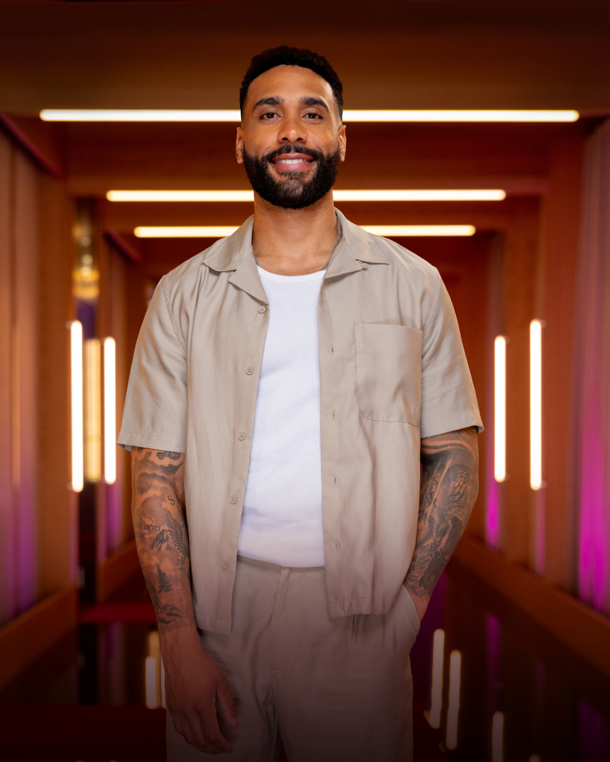 Man with tattoos wearing a light beige short-sleeve shirt and pants stands smiling in a modern hallway with warm lighting, vertical glowing lights, and purple accents in the background.