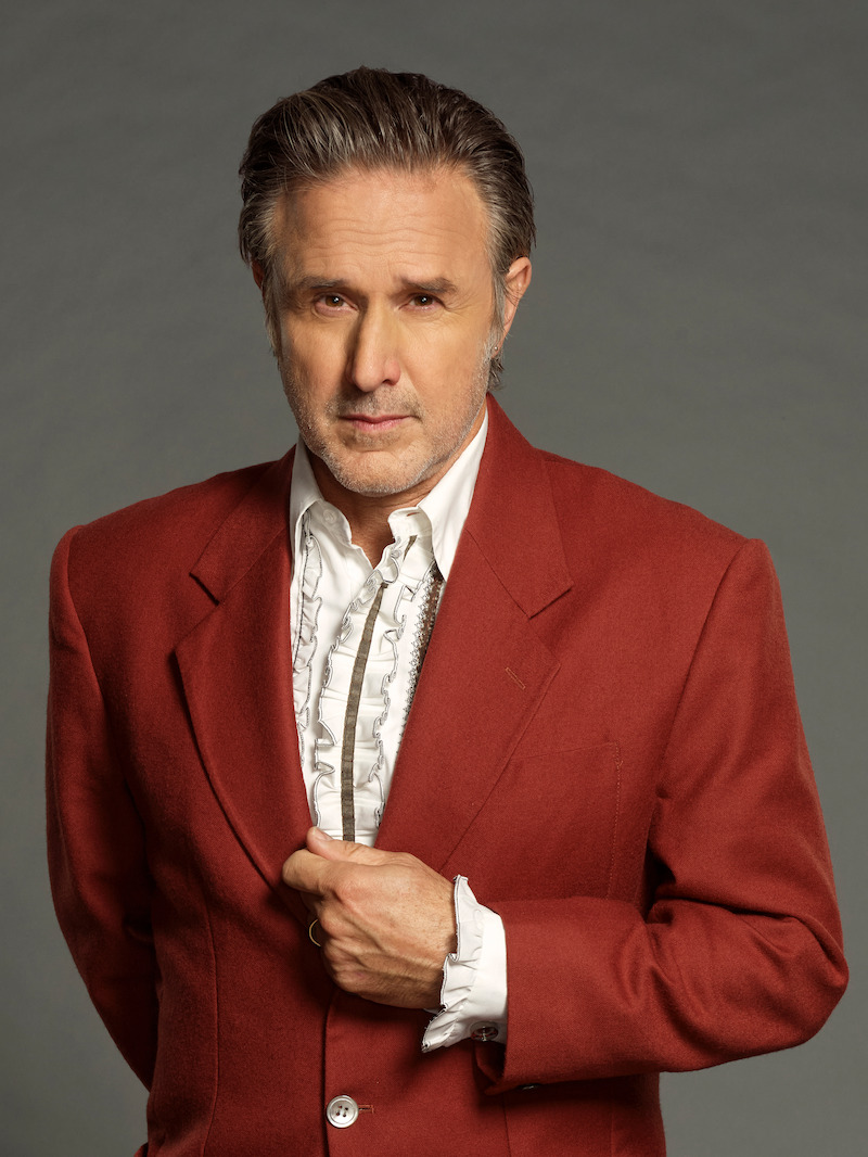 Man with gray hair wearing a red blazer and white ruffled shirt stands against a plain gray background, looking directly at the camera with a serious expression, hand adjusting his jacket.