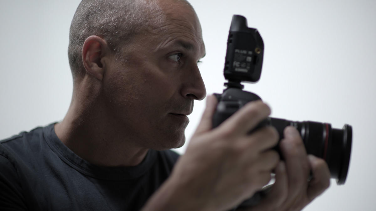 A bald man in a dark shirt holding a camera with a flash attachment, focusing intently in a well-lit, minimal studio environment with a plain light background.