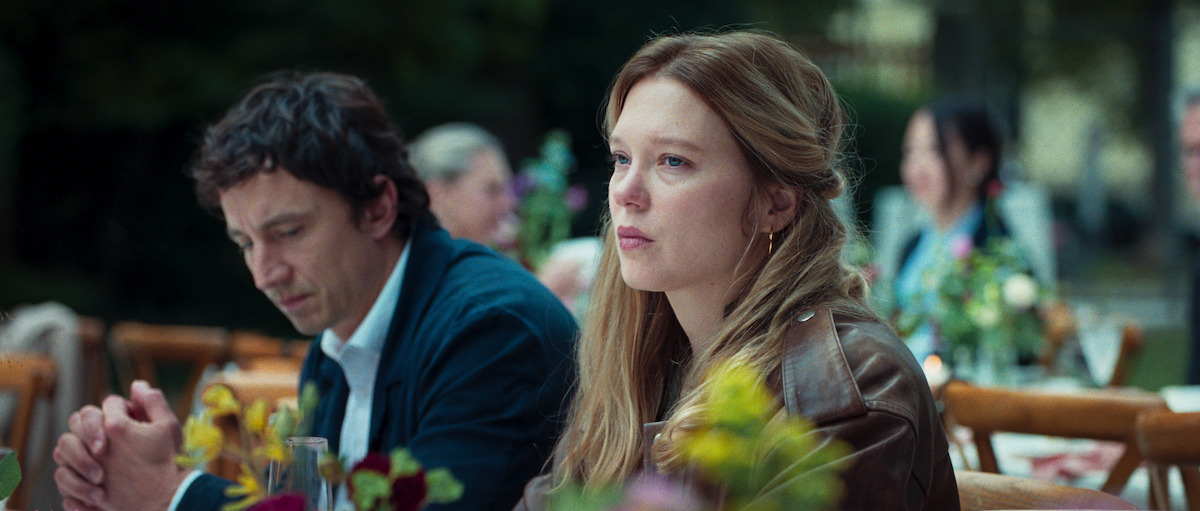 A woman and a man sit outdoors at a table decorated with flowers, surrounded by other people in a garden or park setting, looking thoughtful or pensive.