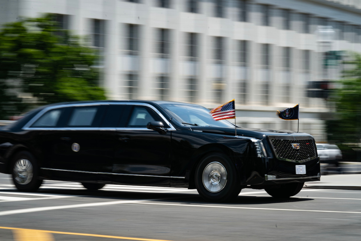 Black presidential limousine with US and official flags drives quickly past a white government building in a city. The image conveys motion and urgency in an official, urban environment.