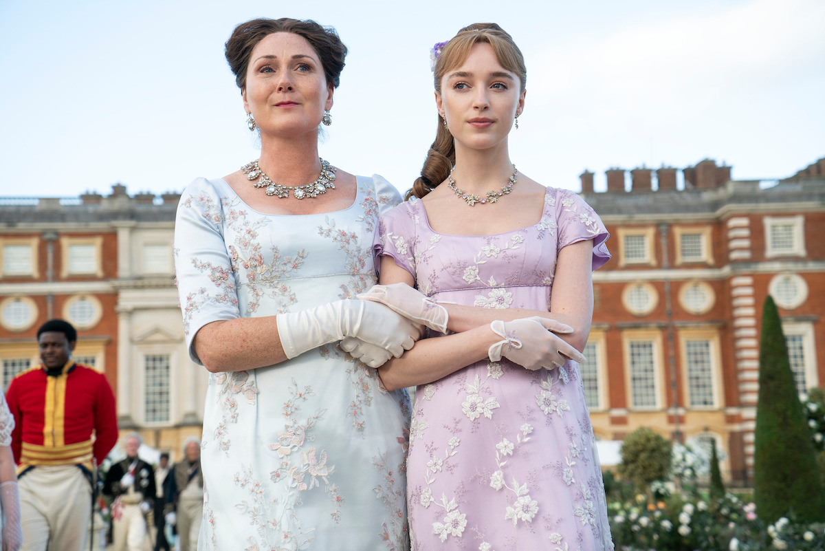 Two women in elegant, Regency-era dresses stand arm-in-arm in a garden, with a grand historic building in the background.